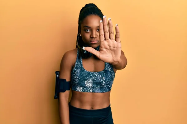 African american woman with braided hair wearing sportswear and arm band doing stop sing with palm of the hand. warning expression with negative and serious gesture on the face.