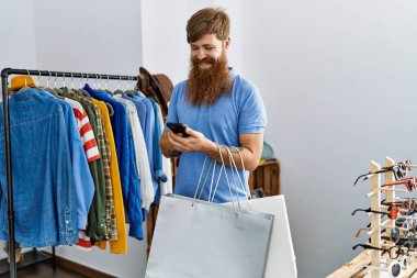 Genç kızıl saçlı müşteri torbaları tutuyor ve giyim mağazasında akıllı telefon kullanıyor..