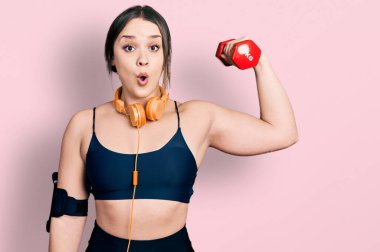 Spor kıyafetleri giyen genç İspanyol kız dambıl kullanıyor. Korkmuş ve ağzı açık şaşkınlık içinde, inançsız bir yüz. 