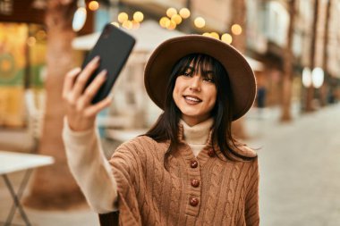 Kışlık şapkalı esmer bir kadın şehirde akıllı telefonuyla selfie çekiyor.