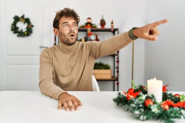 Masada sakallı genç yakışıklı adam Noel süslemeleri ile parmakla göstererek şaşırmış, ağzı açık şaşkınlıkla bakan, ön tarafta bir şey 