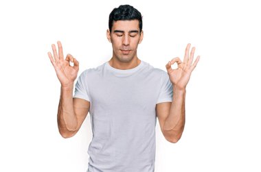 Beyaz tişört giyen yakışıklı İspanyol adam rahatla ve gözleri kapalı gülümse parmaklarıyla meditasyon hareketi yapıyor. yoga konsepti. 