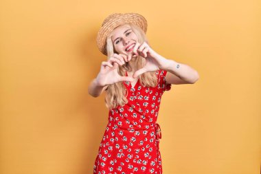 Sarışın, yaz şapkası takan, aşka gülümseyen, elleri ile kalp sembolü yapan güzel beyaz bir kadın. romantik kavram. 