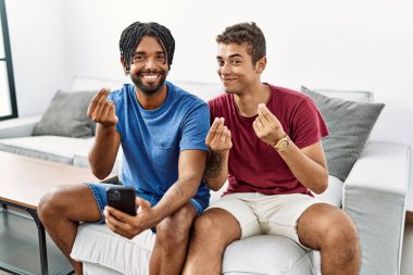Genç ispanyol adamlar akıllı telefon kullanıyor. Evdeki kanepede oturuyor. Elleriyle para jestleri yapıyor. 