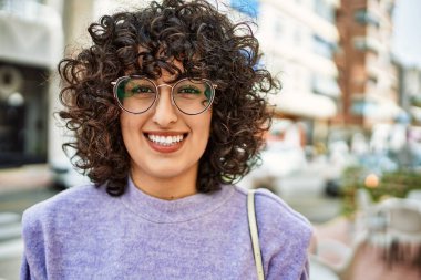 Orta Doğulu genç bir kadın gülümsüyor. Sokakta gözlük takıyor.