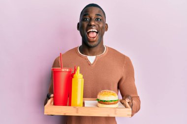 Genç Afrikalı Amerikalı bir adam lezzetli bir hamburger ve soda yiyor çılgın ve başarıyı kutluyor gözleri açık heyecanla haykırıyor. 
