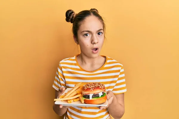 美しいブルネットの女の子はショック顔でフライドポテトとおいしい古典的なハンバーガーを食べて 懐疑的で皮肉 オープン口で驚いています — ストック写真