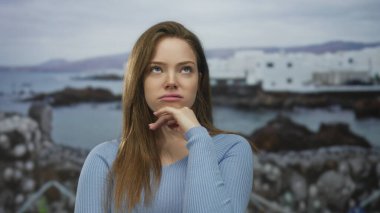 Genç bir kadın, kayalık sahile ve dalgalı suya tepeden bakan beyaz sahil binasının önünde çene altında dinleniyor..