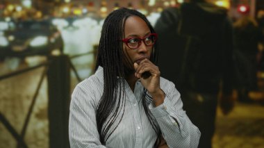 Gözlüklü bir kadın, arka planda bulanık ışıklar ve bokeh etkisi olan bir şehir caddesinde düşünceli bir şekilde duruyor..