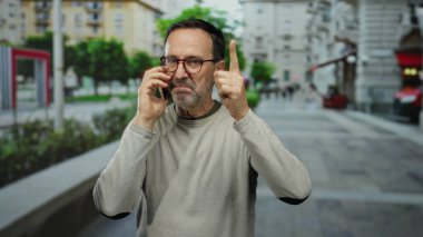 Gözlüklü olgun bir adam, şehir binaları ve yeşilliklerle çevrili akıllı telefonuyla düşünürken bir jest yapıyor..