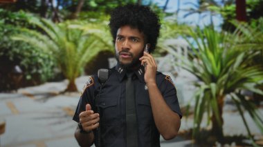 Afro-Amerikan polisi yeşil park ortamında akıllı telefondan konuşuyor. İletişim ve otorite odaklı üniforma giyiyor..