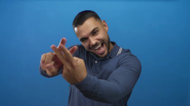 Adam hashtag jesti yapar mavi stüdyo ortamında birbirine dolanmış parmaklarıyla, gülümseyerek ve kameraya doğru eğilerek; neşe ve eğlence iletişimi.