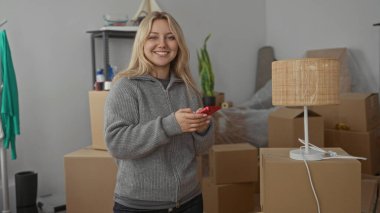 Karton kutular ve ev bitkileriyle çevrili yeni döşenmiş odada akıllı telefona bakarken gülümseyen bir kadın, yakın zamanda rahat bir daireye taşındığını ima ediyor..