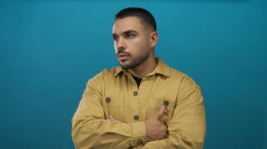 İspanyol adam canlı mavi stüdyo ortamında kollarını çaprazlıyor. Hardal rengi ceket giyiyor. Yanlara bakıyor..
