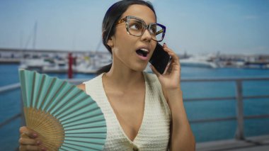 İspanyol kadın telefonda elinde yelpaze, sahil limanında tekneleri ve açık gökyüzü arka planıyla konuşuyor..