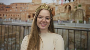 Eski bir altın taç giyen genç beyaz kadın bir binada gözleri ile gülümsüyor..