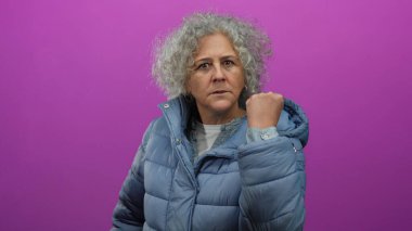 Gri saçlı kadın pembe arka planda mavi ceket giyiyor. Güç ve dayanıklılığı tasvir ederken yumruğunu sıkarak azmini gösteriyor.