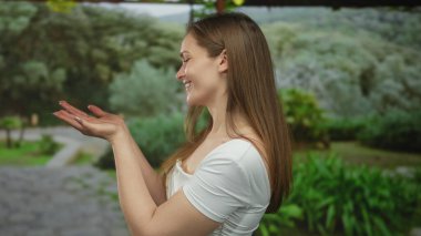 Beyaz tişörtlü bir kadın avuçlarını kaldırıyor ve güneşli bir ormanda gülümsüyor. Çıplak kollarıyla ve rahat duruşuyla, mutluluğu karşılıyor..