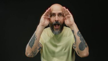 İzole edilmiş siyah bir arkaplan, sakallı, kel kafalı ve nötr renkli tişörtlü bir adama karşı şaşırmış pozlar veren adam belirgin özellikleri vurguluyor..