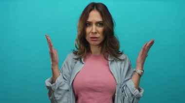Şaşkın bakışlı bir kadın pembe gömlek ve kot ceket giyen, kafa karışıklığı ve merakı taşıyan canlı mavi bir arka plana el kaldırıyor..