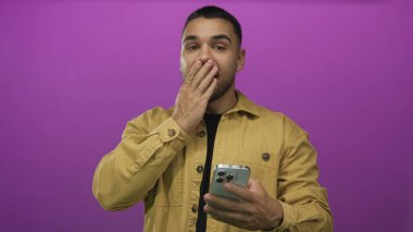 Genç İspanyol adam mor stüdyo ortamında akıllı telefonu tutarken ağzını eliyle kapatıyor. Sürpriz..