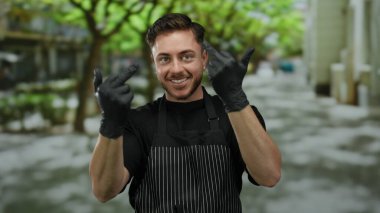 Şehir sokaklarında sakallı genç bir adam önlük ve eldiven giyip orta parmaklarıyla kaba bir jest yapıyor..