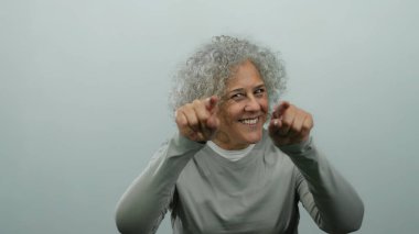 Gri saçlı bir kadın iki eliyle buraya gelir izole edilmiş beyaz bir arka plan duvarına gülümser, dostça bir davetkar tavır sergiler..