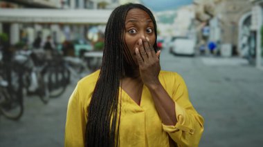 Sarı tişörtlü kıvırcık saçlı kadın, şehir sokaklarında gün içinde bulanık şehir manzarasına sahip şaşırmış yüz ifadesi sergiliyor.