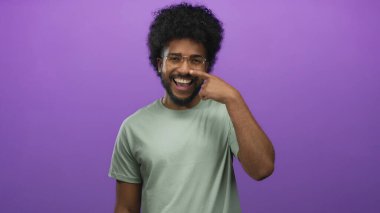 Burnunu gösteren gözlüklü gülümseyen bir adam canlı mor bir arka plana karşı poz veriyor, izole edilmiş bir stüdyoda neşeli ve kendinden emin bir ruh hali sergiliyor..