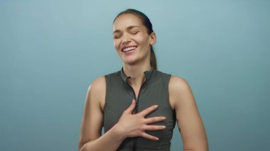Açık mavi bir stüdyo duvarına karşı kolsuz bluz giyen bir kadın göğsüne bastırılmış şekilde gülüyor..