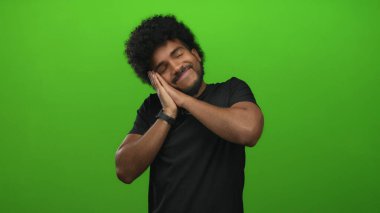 Canlı yeşil bir arka planda uyuyormuş gibi yapan adam, gevşemeyi ve yüzün yanındaki ellerden memnuniyeti ifade ediyor, günlük siyah giysiler giyiyor..