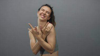 Gri stüdyodaki İspanyol genç kadın elleri ve dili açık rock kornaları gösteriyor; eğlence enerjisi coşkusu.