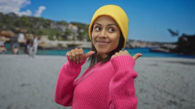 Parlak pembe kazaklı genç İspanyol kadın deniz kenarında neşeyle gülümseyen sarı bir bere takan parlak mavi gökyüzü ile kumsalda geriye doğru hareket ediyor.
