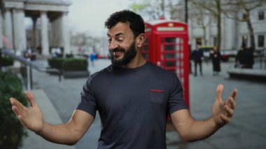 Sakallı İspanyol bir adam klasik İngiliz telefon kulübesi ve arka planda bulanık yayalar olan bir caddenin önünde durup ikonik bir şehir manzarası yakalıyor..