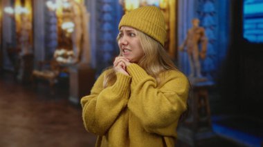 Süslü kilisenin içinde dua eden kadın sarı örgü şapka ve süveter giyiyor ve iç mekanda endişeli bir ifade takınıyor.