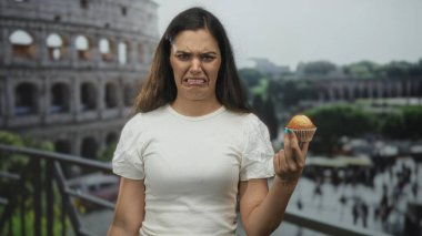 Genç İspanyol kadın, taş binanın yanında kek tutarken kaşlarını çatar ve baş parmağını kaldırır; iğrenme.