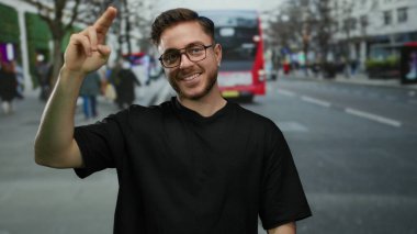 Sakallı genç bir adam, arka planda kırmızı bir otobüsle kalabalık bir şehir sokağında selamlama hareketi yapıyor, şehir yaşamını yakalıyor..