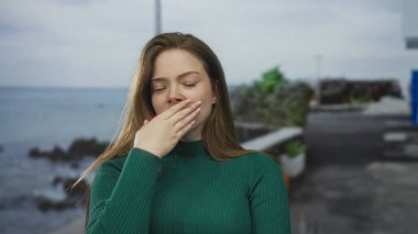 Mavi okyanus dalgaları üzerinde saksılar ve kıyı kayaları ile kaplı bir sahil caddesinde kadın elini ağızla kapatır; yorgunluk.