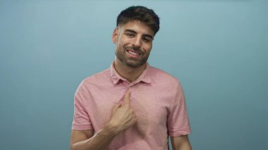 Latin kökenli genç adam gülümser ve açık mavi bir stüdyo ortamında başparmak kaldırır; güven pozitifliği başarı teşviki.