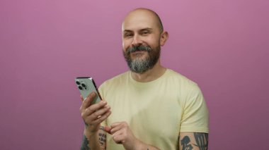 Sakallı ve dövmeli bir adam, modern teknoloji ve iletişimi sembolize eden pembe bir arka plan üzerinde akıllı telefon kullanıyor.