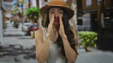 Hasır şapkalı genç kadın elleri ağzında güneşli bir şehir caddesinde bağırıyor; acil çağrı.