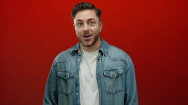 Kot gömlekli sakallı genç adam canlı kırmızı arka planda dalgın dalgın bakıyor.