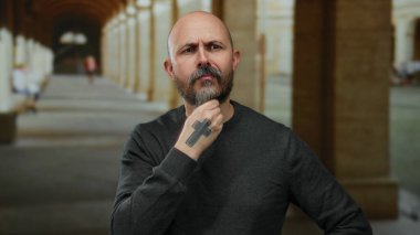 Dövmeli kel bir adam. Üniversite koridorunda el yordamıyla düşünüyor. Düşünceli bir yüz ifadesi ve arka planda kemerleri olan sakalı var..