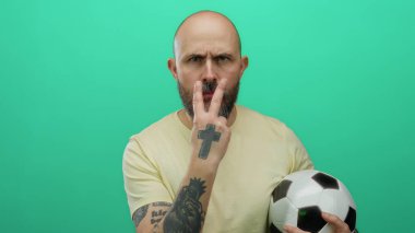 Futbol topu tutan sakallı kel bir adam, izole edilmiş yeşil arka plana karşı tehditkar hareketler yapıyor, duygularını ve yoğunluğunu açıkça ifade ediyor..