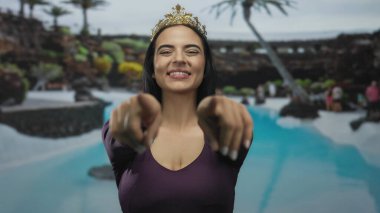 Taç giymiş bir kadın havuz kenarında kameraya gülümsüyor. Etrafı yemyeşil bir otel ortamında tatil havası ve rahatlama hissi veriyor..