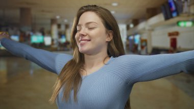 Mavi kazak giyen bir kadın havaalanı kontrol noktasında kollarını kaldırıp gülümsüyor..