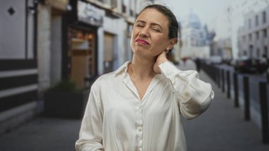 Bulanık mağazaların ve park edilmiş arabaların ortasında, şehir caddesinde beyaz gömlekli bir kadın boynuna dokunuyor..
