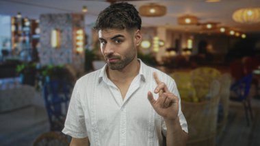 Sakallı bir adam, geniş bir otel binasında beyaz keten bir gömlek giyerken parmağını gösteriyor..