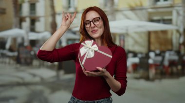 Terasta hediyeyle gülümseyen, gözlüklü ve kırmızı gömlekli bir kadın şehir restoranında neşe saçıyor..