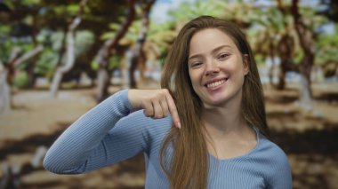 Mavi tişörtlü genç kadın gülümsüyor ve parmakla işaret ediyor güneş ışığı altındaki yeşil orman ağaçlarının arasında; mutluluk.